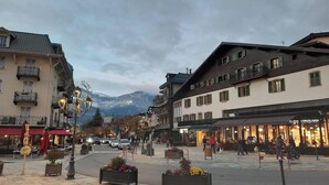 Une place de ville bordée de bâtiments, avec une montagne en arrière-plan et des espaces aménagés pour s’asseoir à l’extérieur.