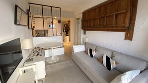 Un salon moderne avec un canapé gris, un téléviseur, une petite table et, au fond, un coin cuisine.