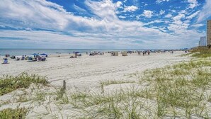 Une plage avec des gens et des parasols, un ciel dégagé et un bâtiment en arrière-plan.