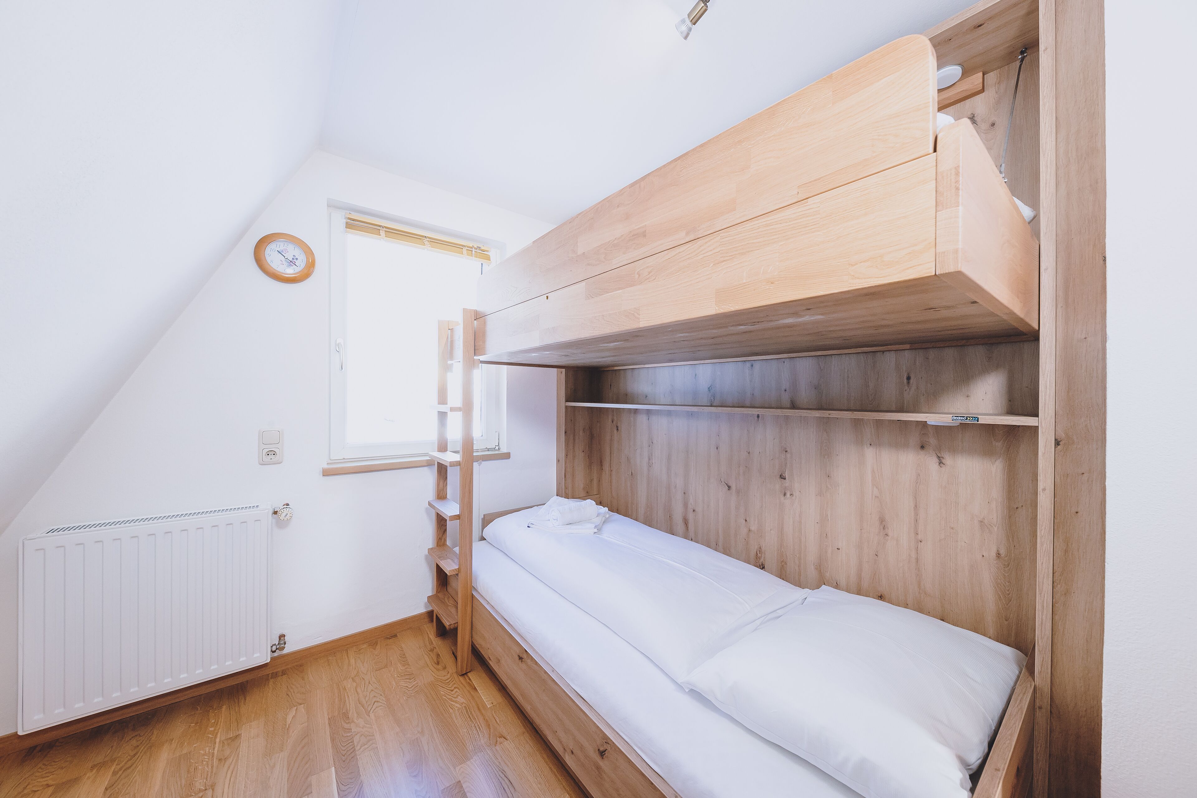 A dorm-style room with a bunk bed, wooden walls, a white radiator, a window with a wooden frame, and a wall clock.