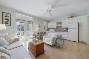 A spacious living room with a sofa, coffee table, and a dining area with chairs. A kitchen with white cabinets and a refrigerator is visible in the background.