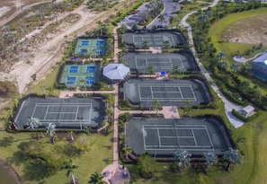Eine Vogelperspektive eines Sportkomplexes mit mehreren Tennisplätzen, einem zentralen Pavillon und umgebender Grünfläche.