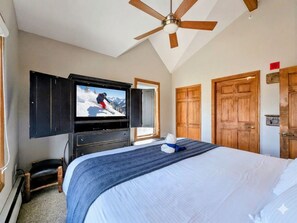 Un dormitorio con una cama, un ventilador de techo, un televisor instalado en la pared y puertas de madera.