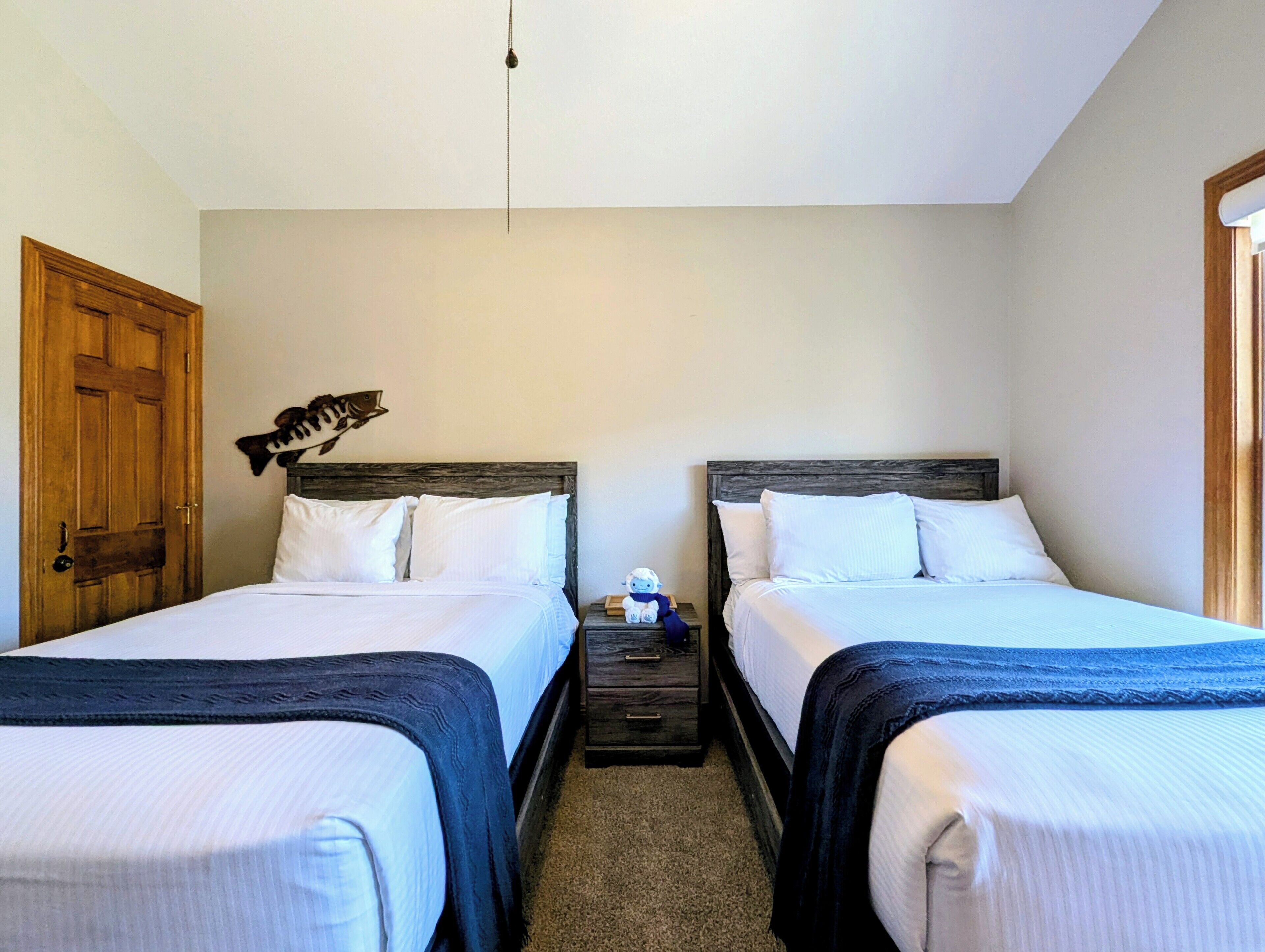 A room with two beds, a wooden door, a wall-mounted fish decoration, and a nightstand with a lamp.