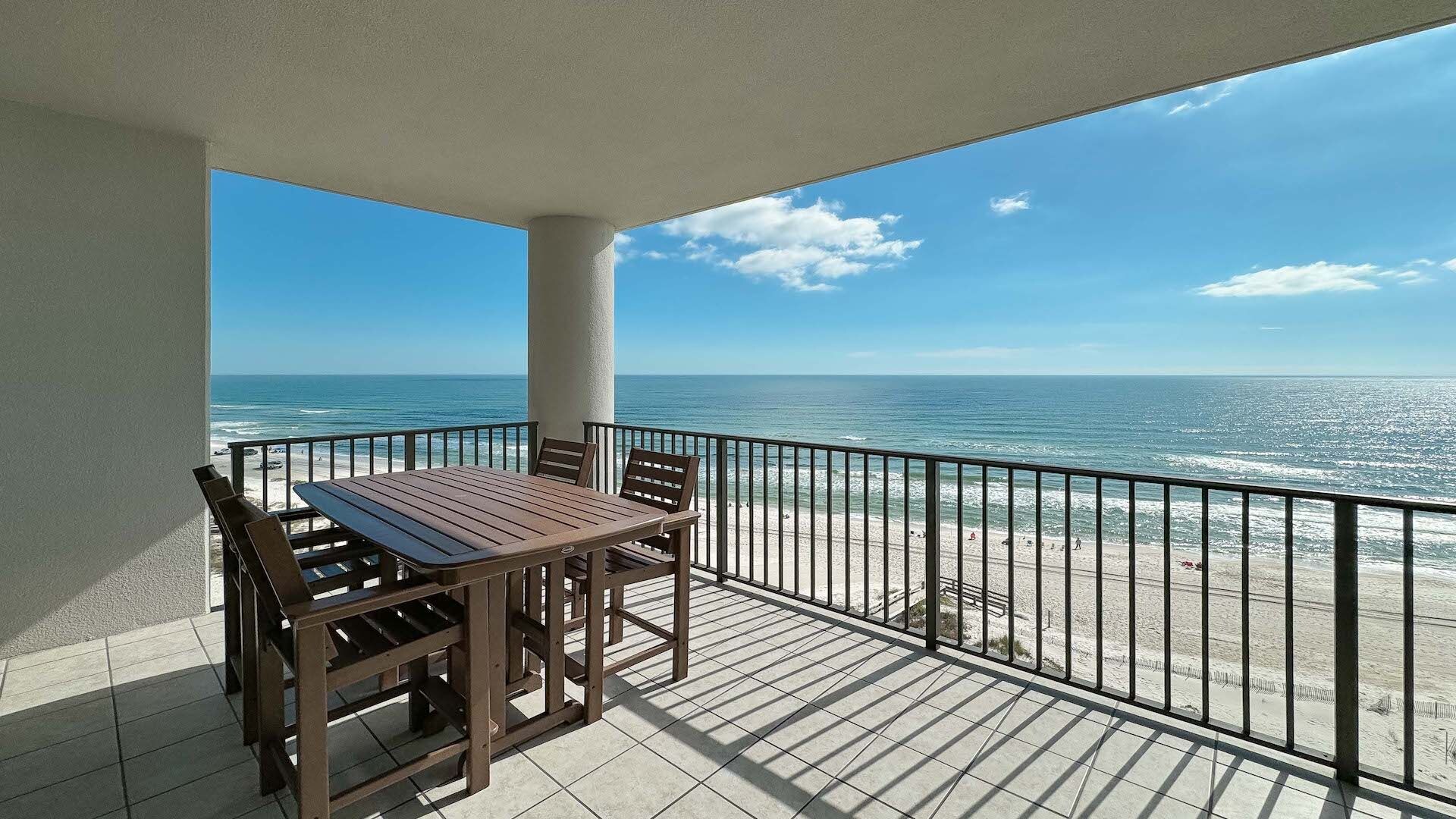 A balcony with a table and chairs, overlooking a beach with the ocean and a clear sky.