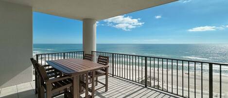 A balcony with a table and chairs, overlooking a beach with the ocean and a clear sky.