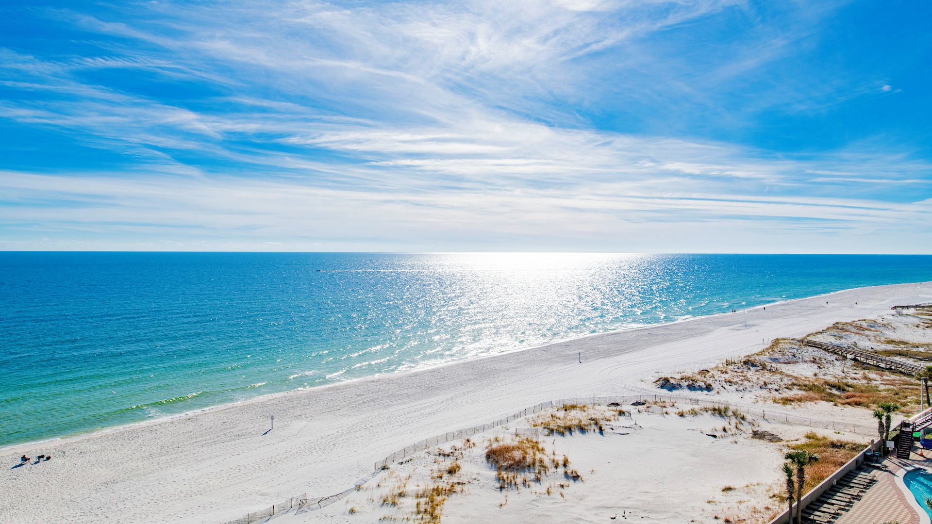 A beach with a clear sky, a sandy shore, and a wooden boardwalk leading to a pool area.