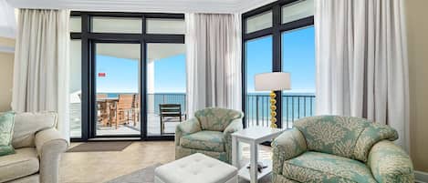 A hotel room with two green patterned sofas, a white ottoman, a small white side table, and a floor lamp. There is a sliding glass door leading to an outdoor area with a view of the ocean.