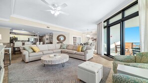 A spacious living room with a large sectional sofa, a round coffee table, and a view of the ocean through large windows. - 7th Floor!!  Recently Redecorated, Lazy River and Slide!! (Orange Beach)