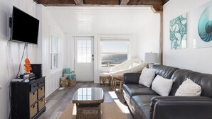 A living room with a black leather sofa, a glass coffee table, wicker furniture, and artwork on the walls. - The Cottage @ Dolphin's Watch Down (Folly Beach)