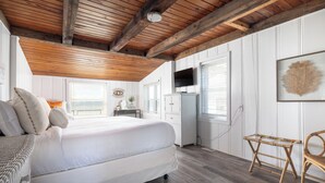 A bedroom with a large bed, wooden ceiling beams, white walls, and a window with blinds.