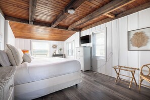 A bedroom with a large bed, wooden ceiling beams, white walls, and a window with blinds.