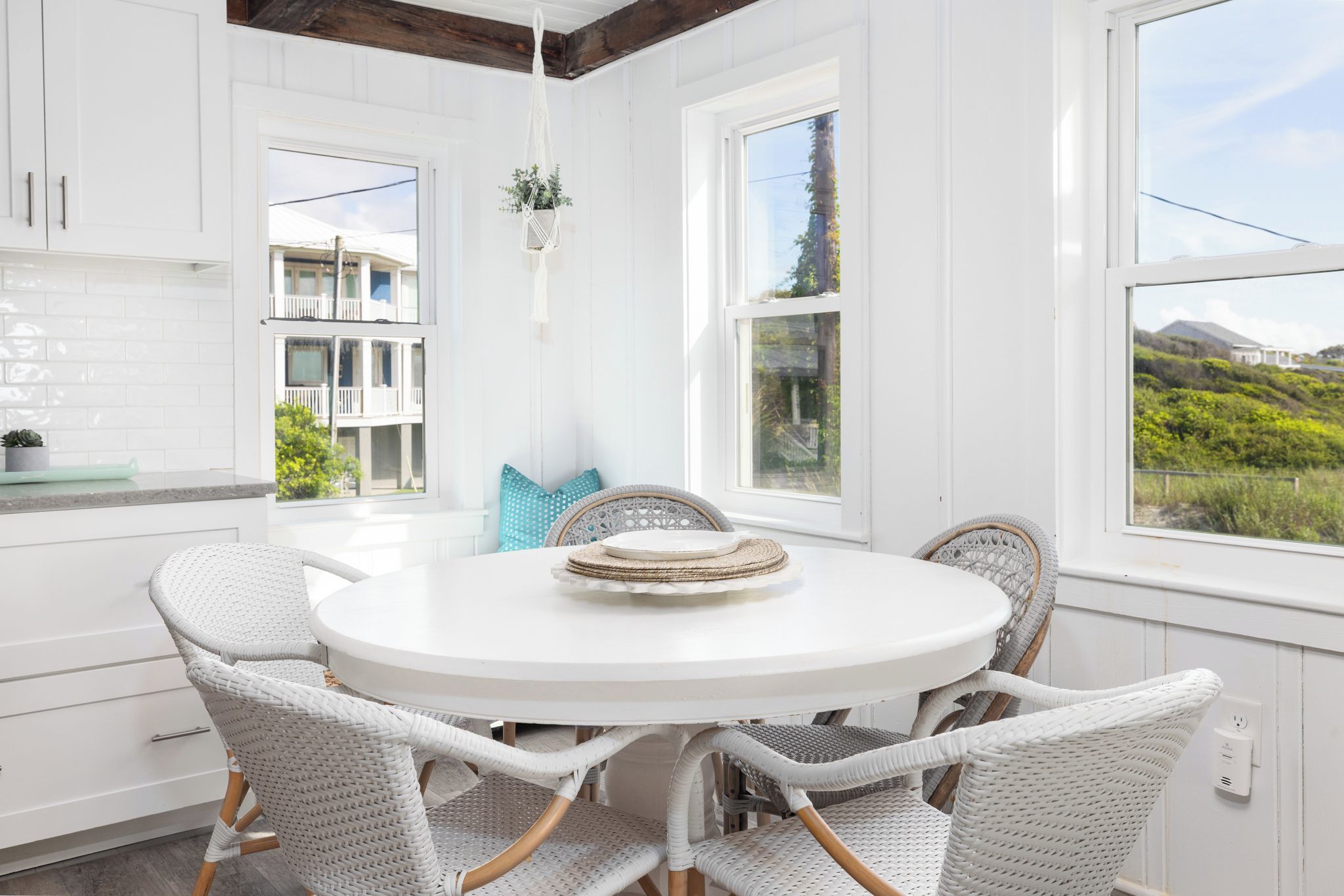Une cuisine blanche avec une table de salle à manger ronde et des chaises en osier, de grandes fenêtres et une plante suspendue.