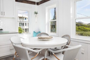 Une cuisine blanche avec une table de salle à manger ronde et des chaises en osier, de grandes fenêtres et une plante suspendue.