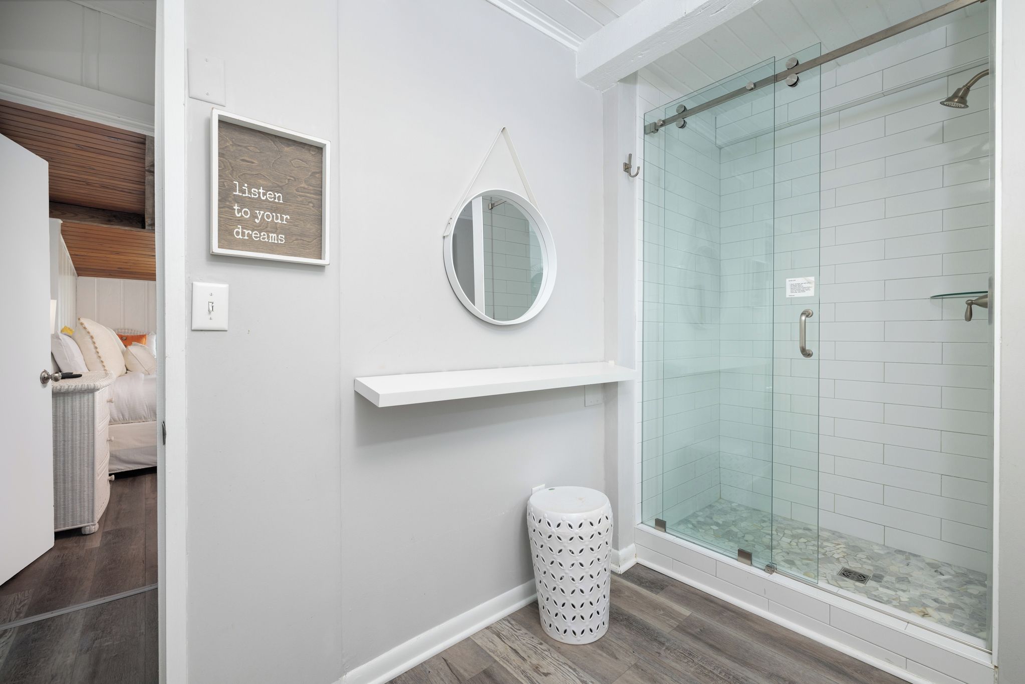 A bathroom with a glass shower enclosure, a round mirror, a framed inspirational quote, and a small trash bin.