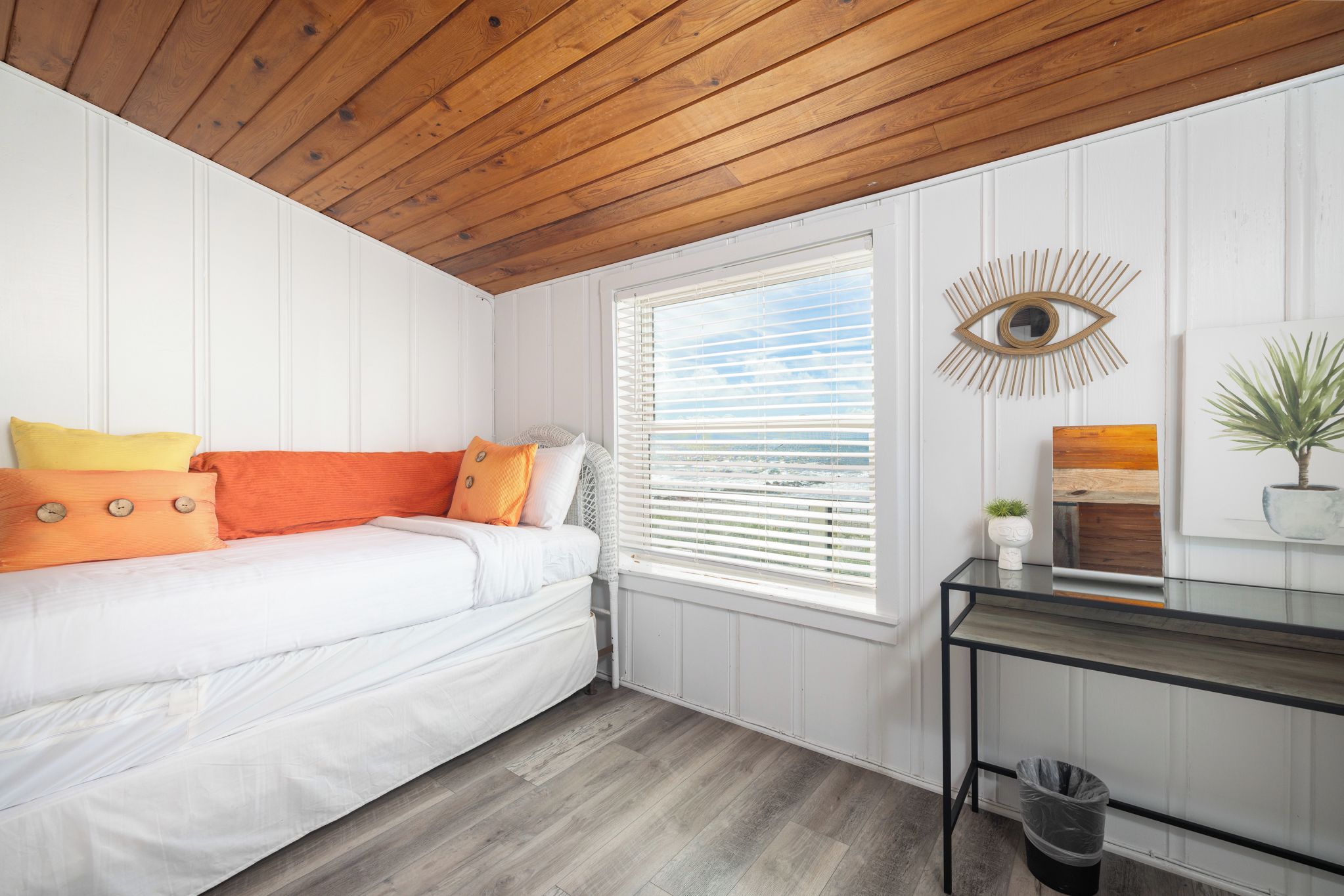 A bedroom with a white bed, orange pillows, a wooden ceiling, a window with blinds, a wall-mounted eye decoration, and a small table with a plant.