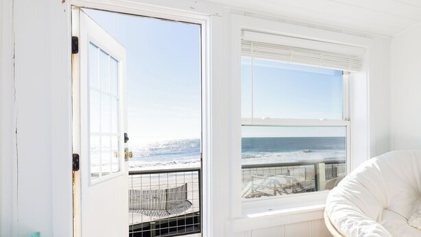 A room with a view of the ocean, a wicker chair, a white sofa, and a wicker basket. - The Cottage @ Dolphin's Watch Down (Folly Beach)