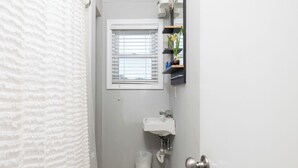Ein Badezimmer mit einem weißen Waschbecken, einem Fenster mit Jalousien, einem Mülleimer und einer Tür.