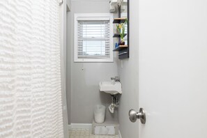 A bathroom with a white sink, a window with blinds, a trash bin, and a door. - "The Retreat" at Dolphin's Watch - Up (Folly Beach)