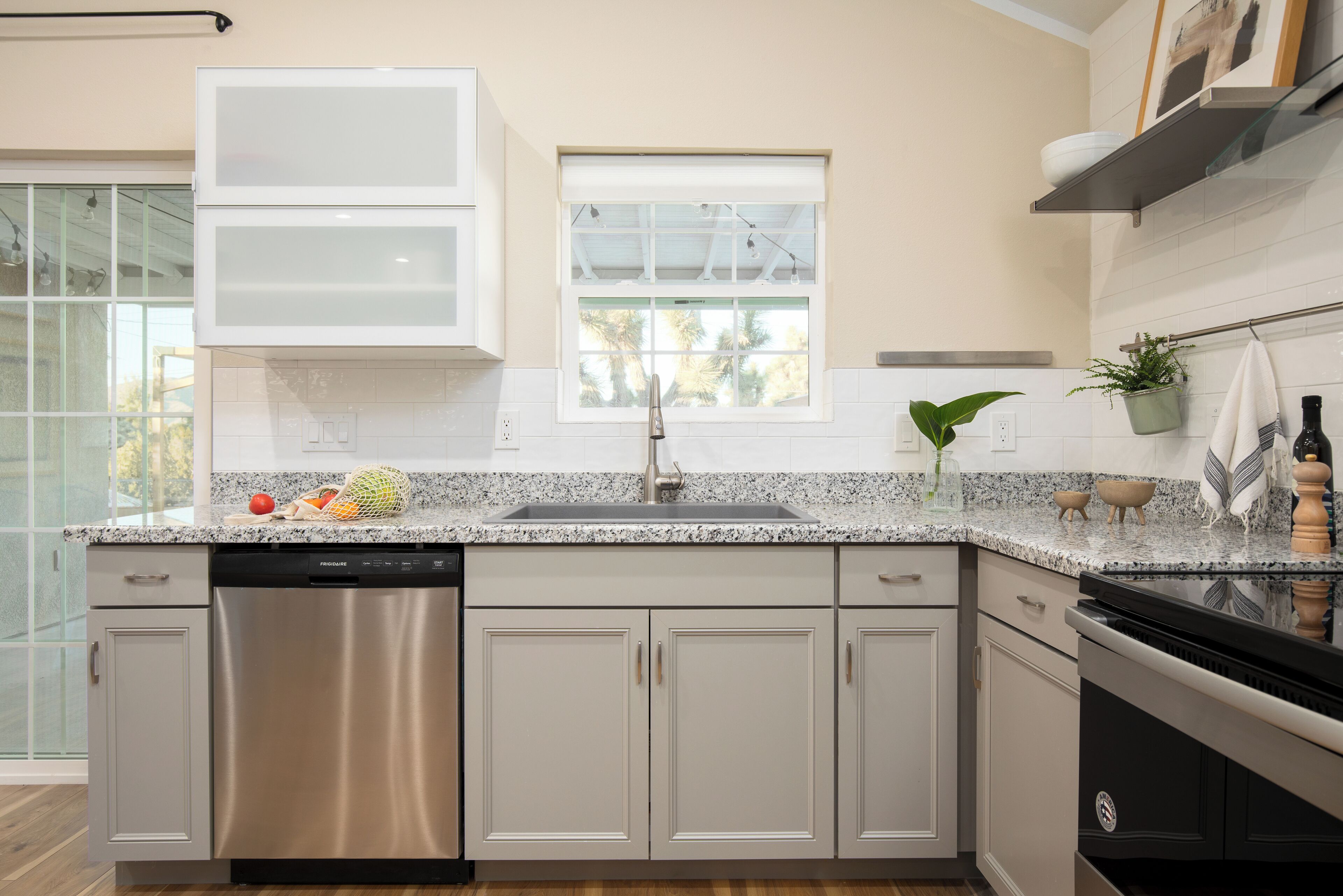 A modern kitchen with a dishwasher, stainless steel cabinets, a granite countertop, and a window with a view of palm trees.