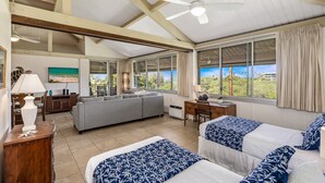 A spacious room with a high ceiling, large windows, a ceiling fan, a sofa, a desk, and two beds with patterned bedding.