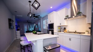 A modern kitchen with white cabinets, a central island with bar stools, a stainless steel range hood, and a ceiling fan.