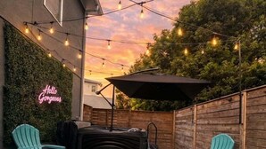 Un patio al aire libre con un alfombra estampada, dos sillas color turquesa, una parrilla para barbacoa y luces colgantes.