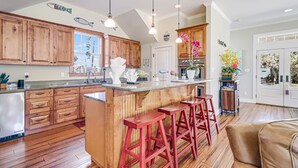 Une cuisine spacieuse avec des meubles en bois, un îlot central doté de tabourets, un éclairage suspendu et une vue sur l’extérieur.