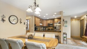 A dining area with a wooden table and chairs, a kitchen in the background, and a chandelier.