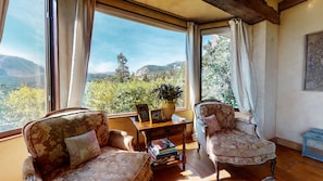 A room with two upholstered chairs, a wooden table, and a view of a mountainous landscape through large windows.