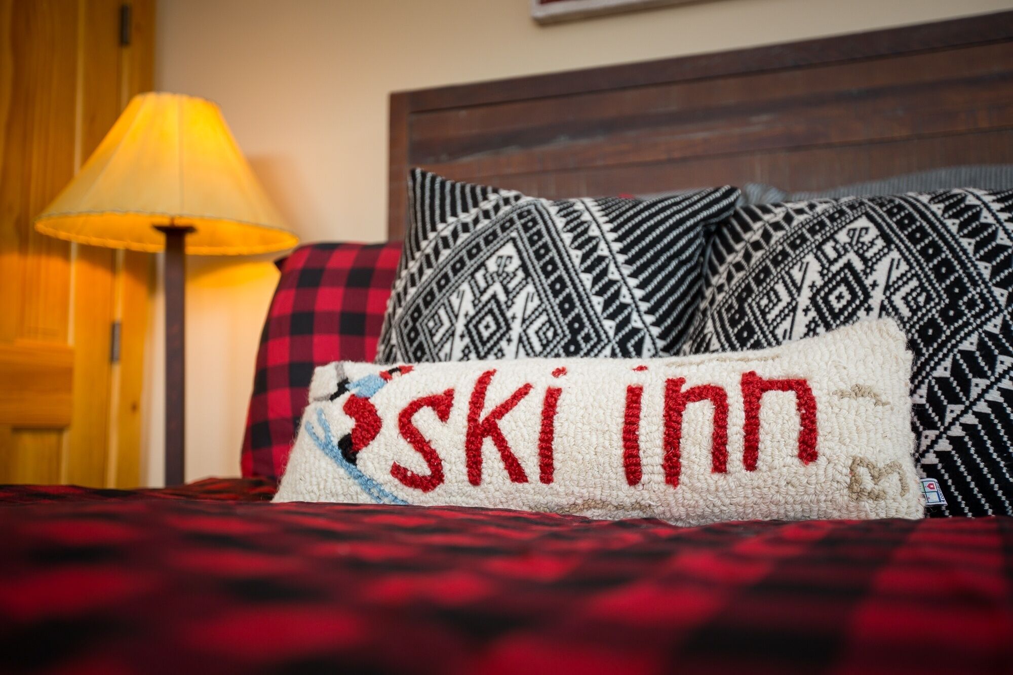 A bed with a red and black checkered blanket, a pillow with 'ski inn' embroidered in red, and a patterned pillow.