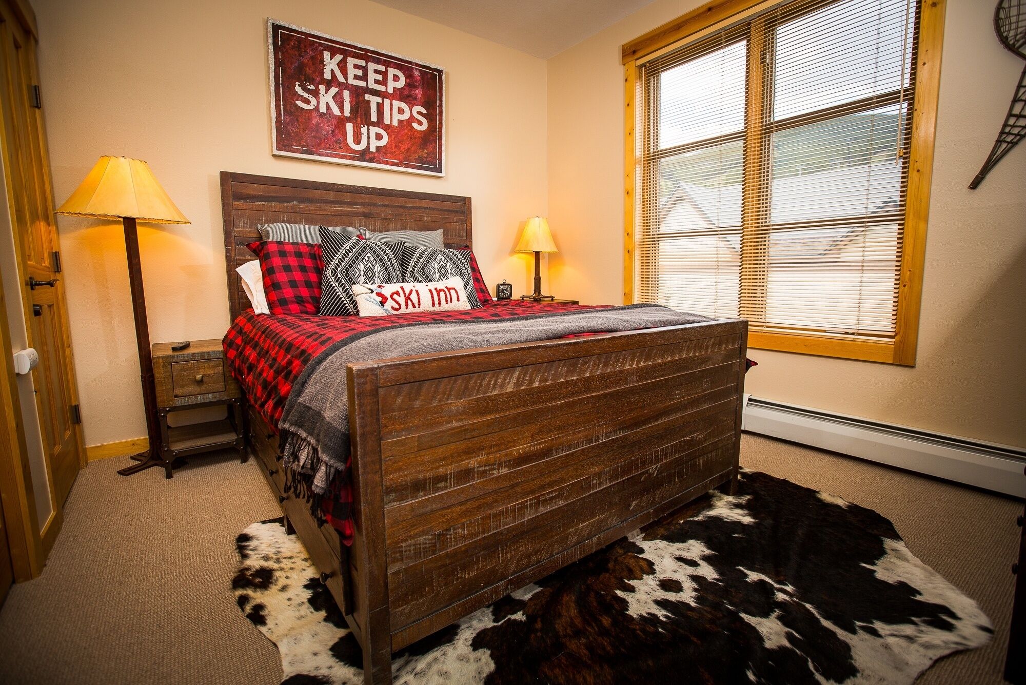 A bedroom with a wooden bed, a cowhide rug, a sign reading "KEEP SKI TIPS UP", and a window with blinds.