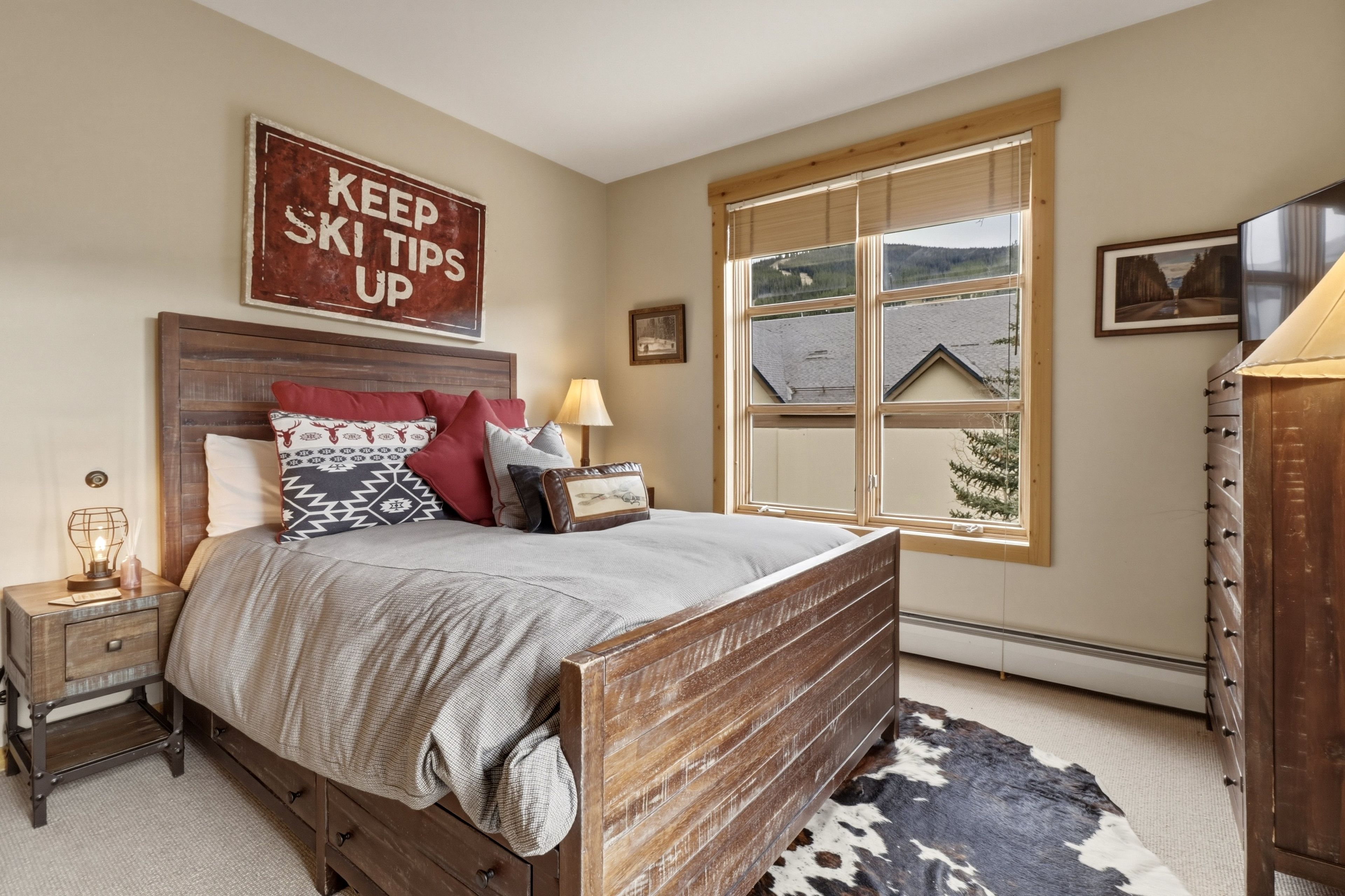 Une chambre à coucher avec un lit en bois, une table de chevet, une lampe, une fenêtre donnant sur une maison et un panneau accroché au mur.