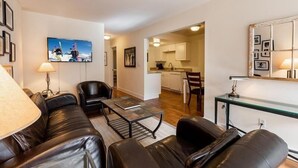Una sala de estar moderna con un sofá de cuero negro, una mesa de centro y un televisor montado en la pared. Se observa una cocina al fondo con armarios blancos y un área de comedor.
