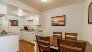 Cocina compacta con armarios blancos, una mesa de comedor con cuatro sillas de madera y dos cuadros en las paredes.