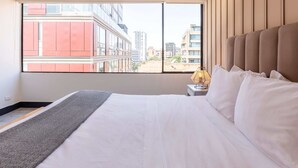 A hotel room with a large window, a bed with white linens, a patterned rug, and a nightstand with a lamp. - w* | Classy 1BR Penthouse in Cabrera (Bogotá)