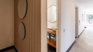 A hallway with a wooden floor, a white wall with a circular mirror, and a door leading to a bathroom with a sink and toilet. - w* | Classy 1BR Penthouse in Cabrera (Bogotá)