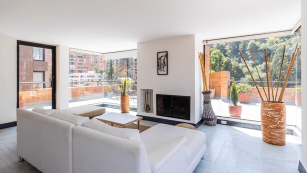 A modern living room with a white sofa, a fireplace, and a view of a cityscape through large windows. - w* | Classy 1BR Penthouse in Cabrera (Bogotá)