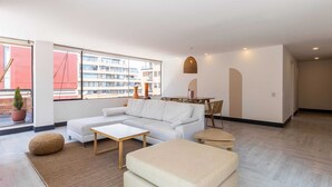A modern living room with a white sofa, a coffee table, and a dining table with chairs. There is a large window with a view of buildings outside. - w* | Classy 1BR Penthouse in Cabrera (Bogotá)