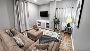 Ein modernes Wohnzimmer mit einem beigen Sofa, einem Couchtisch und einem gemusterten Teppich. An der Wand ist ein Flachbildfernseher angebracht, es gibt eine Zimmerpflanze und einen kleinen Tisch mit einer Lampe.