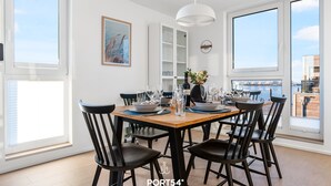 Un coin repas avec une table en bois, des chaises noires et un luminaire suspendu blanc. De grandes fenêtres offrent une vue sur la mer.