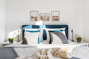 Cama bem arrumada com cabeceira azul, roupa de cama branca e almofadas decorativas. Três quadros emoldurados estão na parede acima da cama.