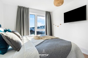 Quarto com cama grande, cortinas cinzentas, televisão na parede e vista para prédios.