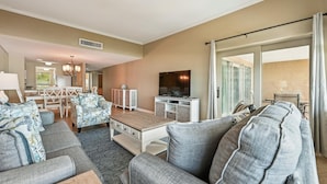 A spacious living room with a large sofa set, a coffee table, a TV, and a dining area with a chandelier. - Coastal 5th Floor Ocean Front Views w/ Amenites (Hilton Head Island)