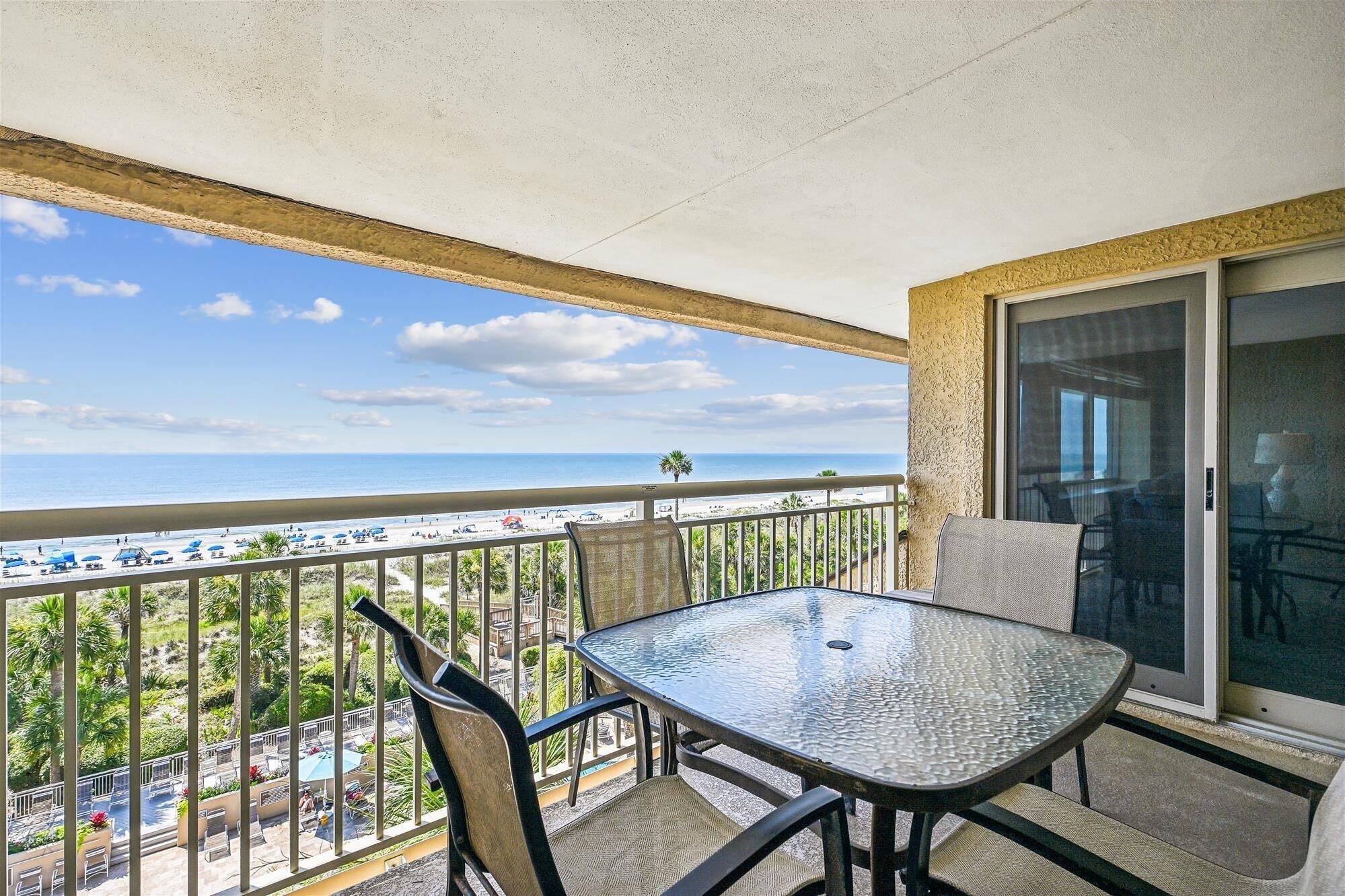 A balcony with a glass table and chairs, overlooking a beach with umbrellas and people.