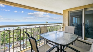 A balcony with a glass table and chairs, overlooking a beach with umbrellas and people. - Coastal 5th Floor Ocean Front Views w/ Amenites (Hilton Head Island)