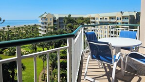 A balcony with a table and chairs overlooking a beach and hotel buildings.