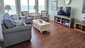 A living room with a large window overlooking the beach, a television, and a bookshelf. - 5th Floor Villa, Steps to the Beach & Downtown (Hilton Head Island)