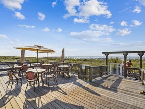 A wooden deck with outdoor seating, including tables and chairs, overlooking a beach with umbrellas and a wooden structure.