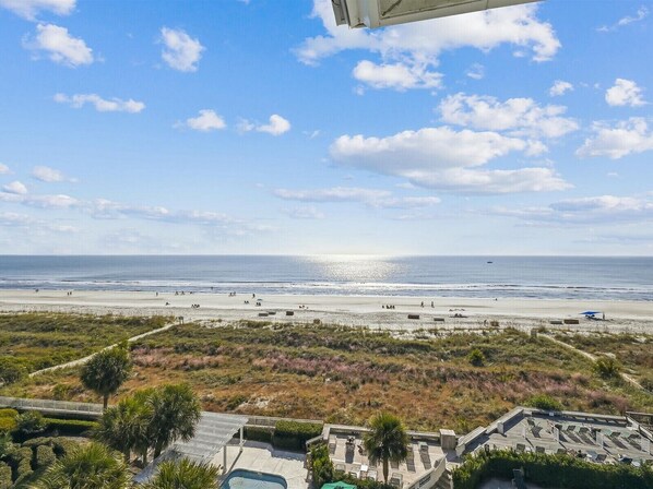 A beach view with a clear sky, a sandy beach, and a few people. There is a structure with a roof and a swimming pool in the foreground.
